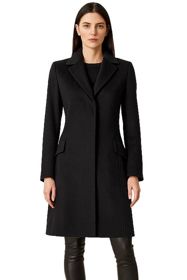 Front view of a person wearing a tailored black knee-length coat with notched lapels, hidden front closure, and flap pockets, paired with black leather pants on a white background.