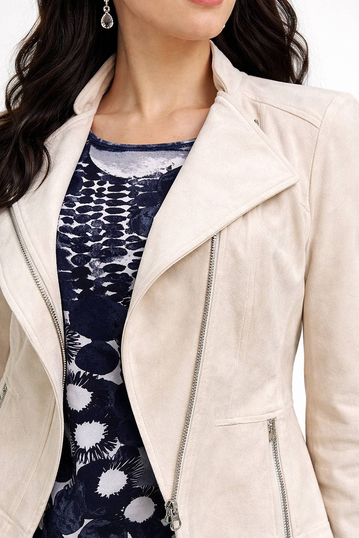 A person is wearing a light beige jacket over a dark patterned shirt, accessorized with earrings.