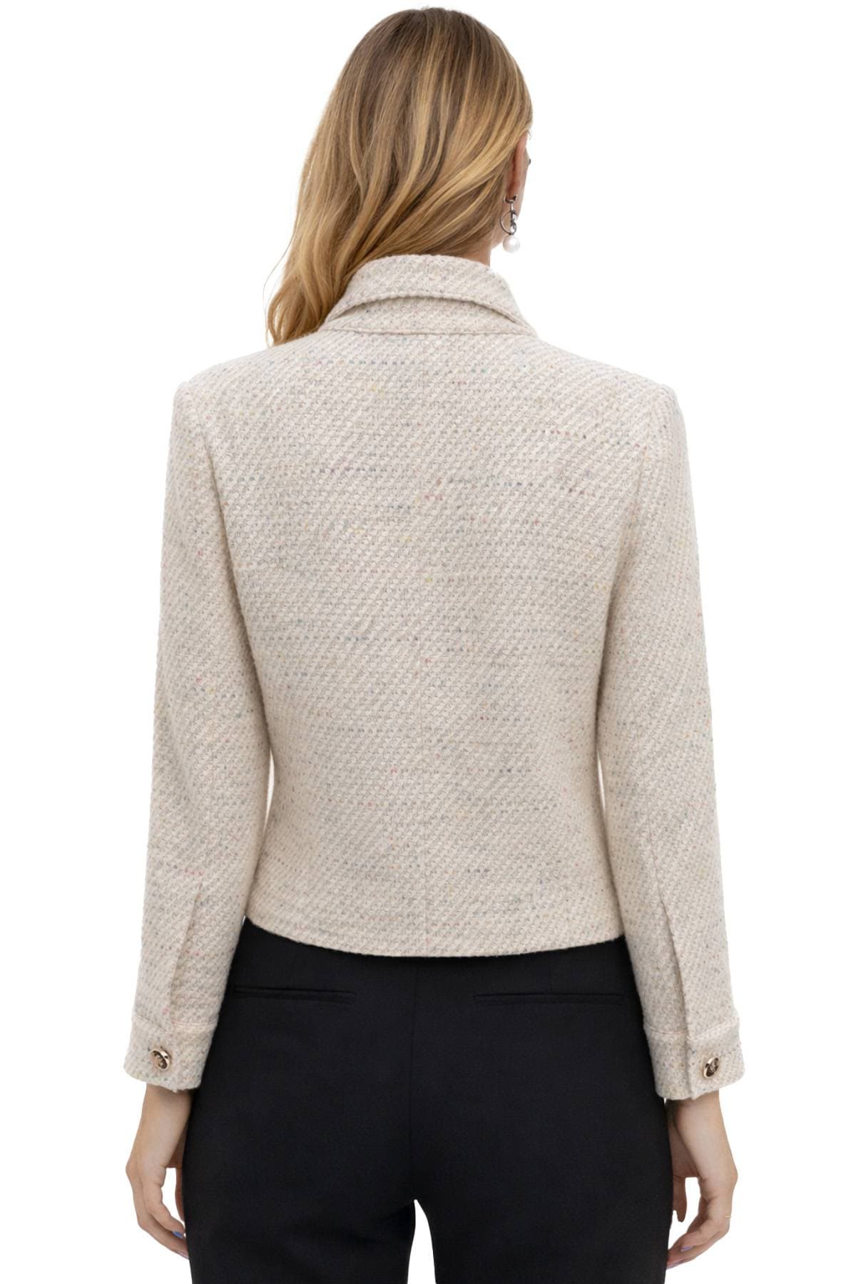 A woman with long hair is wearing a light tweed jacket and black pants, viewed from the back.