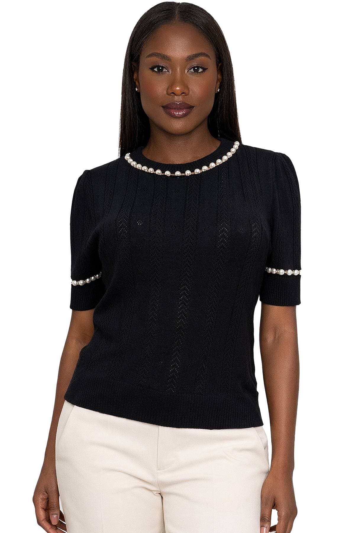 A woman with long hair is wearing a black sweater with pearl details and light-colored pants.