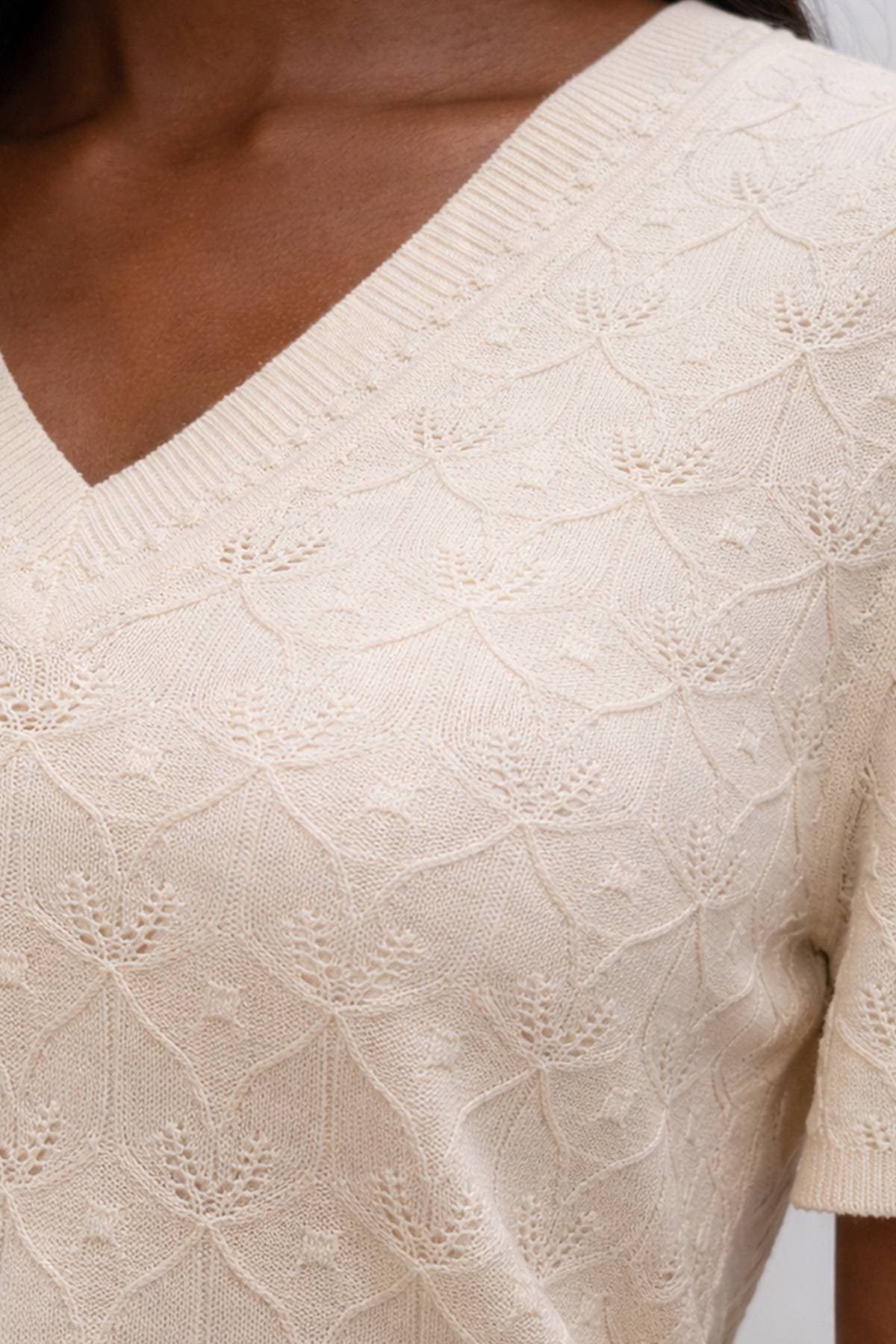 A close-up of a person wearing a cream knit sweater with a V-neck and floral pattern.