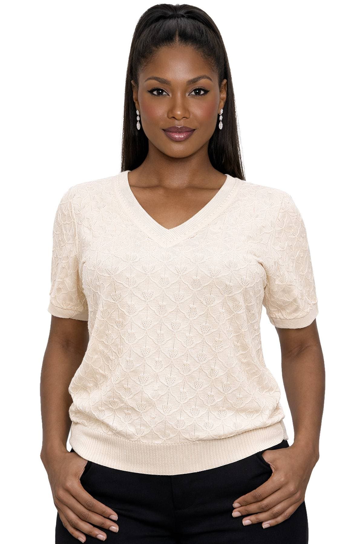 A woman wearing a light beige knit sweater and black pants is looking at the camera with a neutral expression.