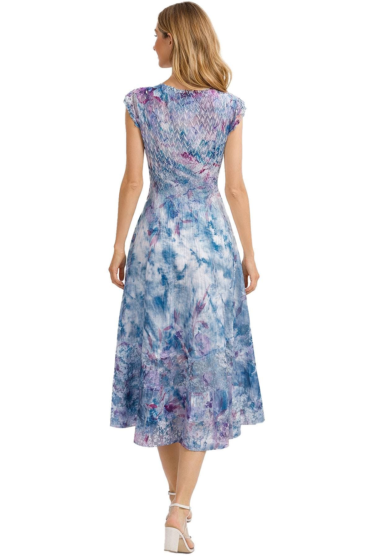 A woman with long blonde hair is wearing a colorful, sleeveless dress with a smocked bodice and a floral pattern, viewed from the back.