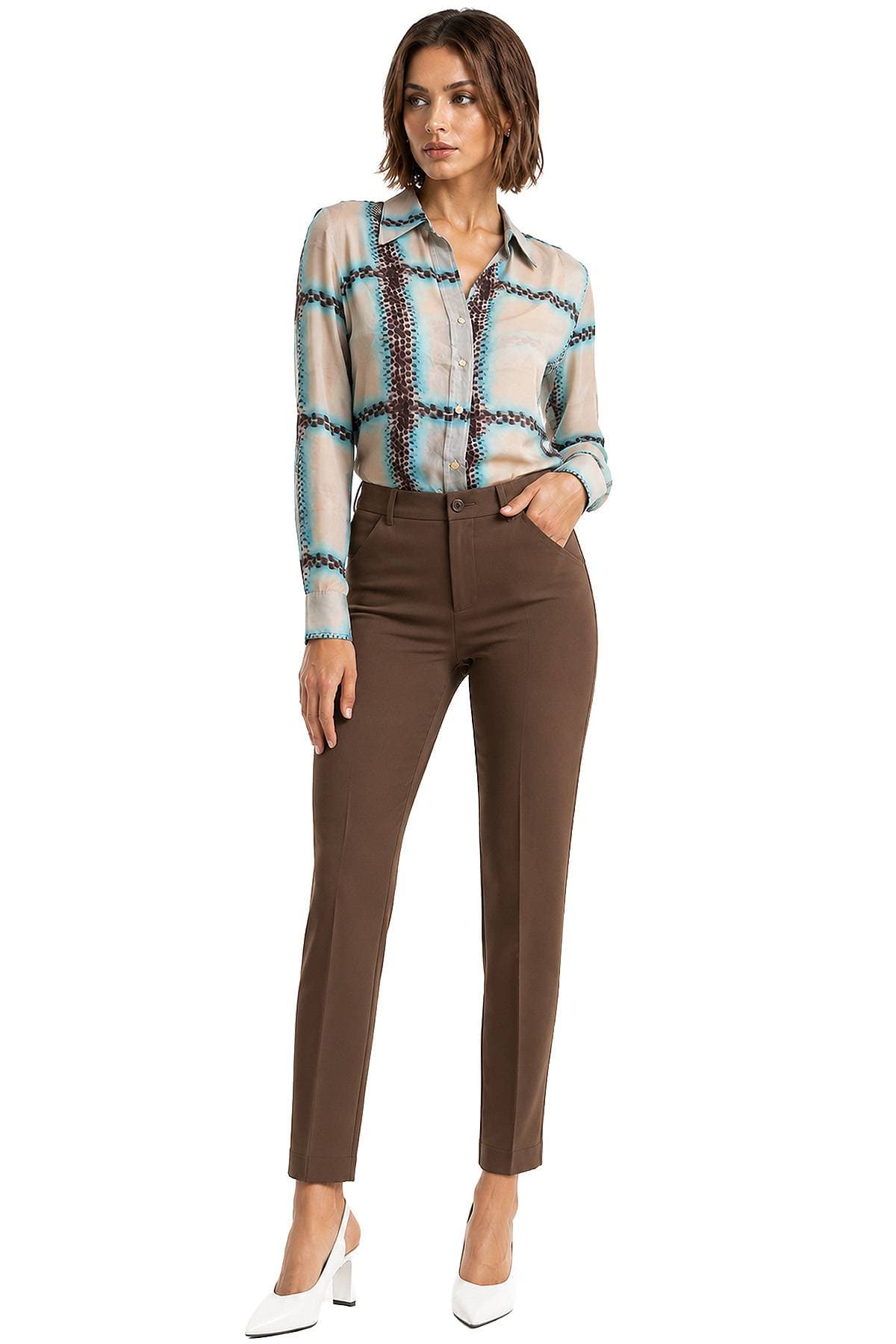 A woman wearing a patterned blouse and brown trousers, posing with one hand in her pocket.