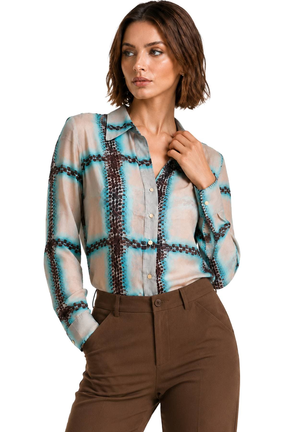 A woman stands wearing a patterned long-sleeved blouse and brown trousers.