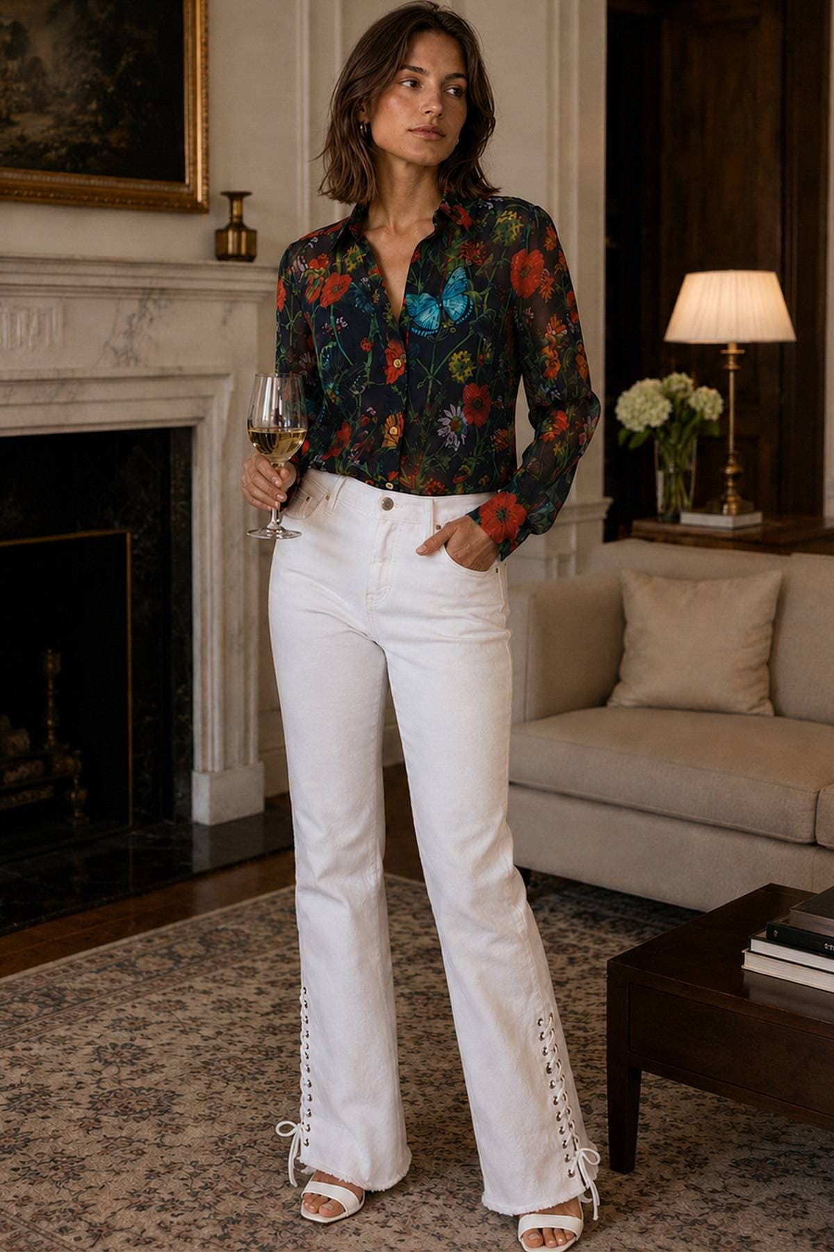 A woman in a floral blouse and white lace-up pants holds a glass of white wine.