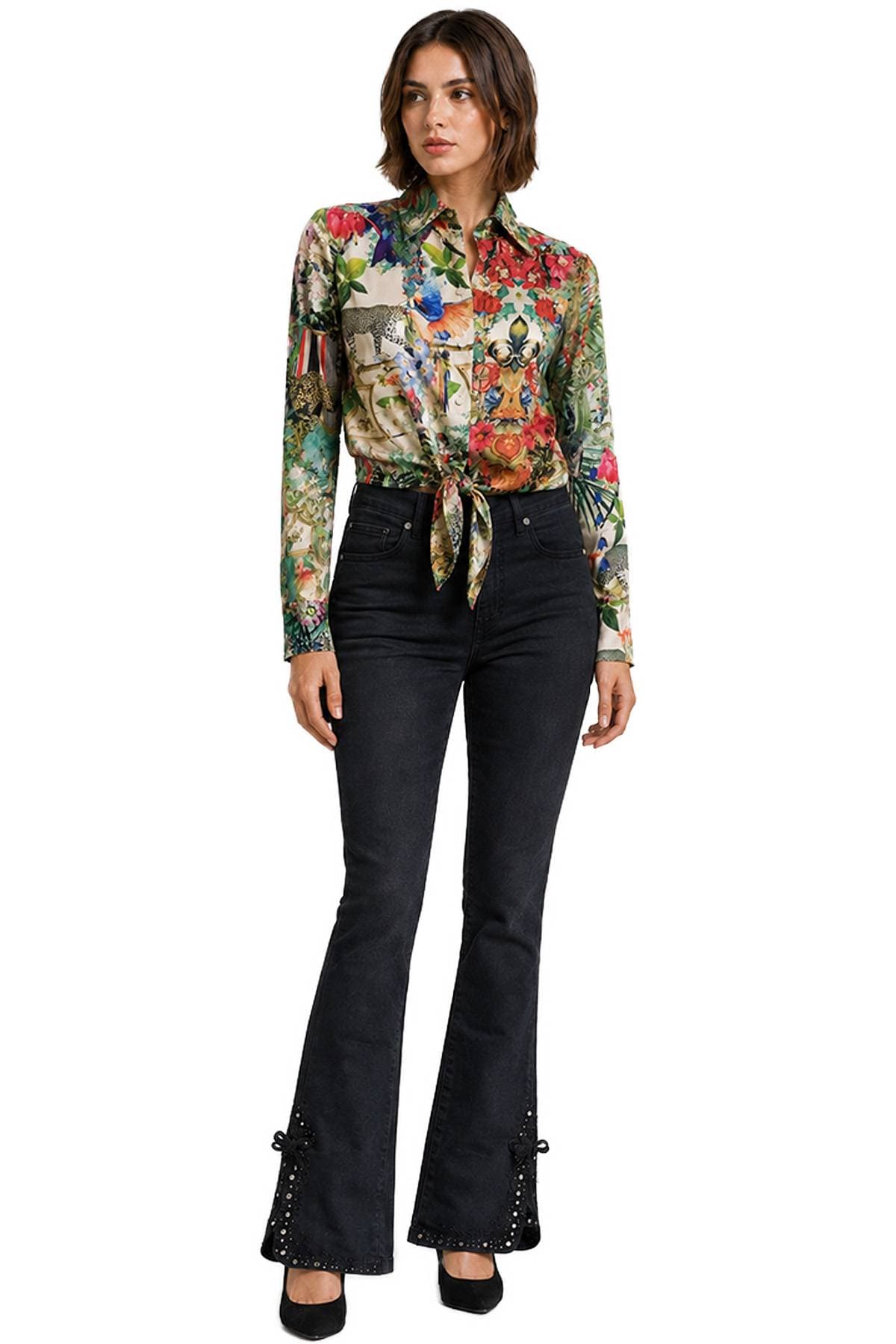 A person wearing a colorful floral shirt tied at the waist and black flared jeans is standing and looking off to the side.