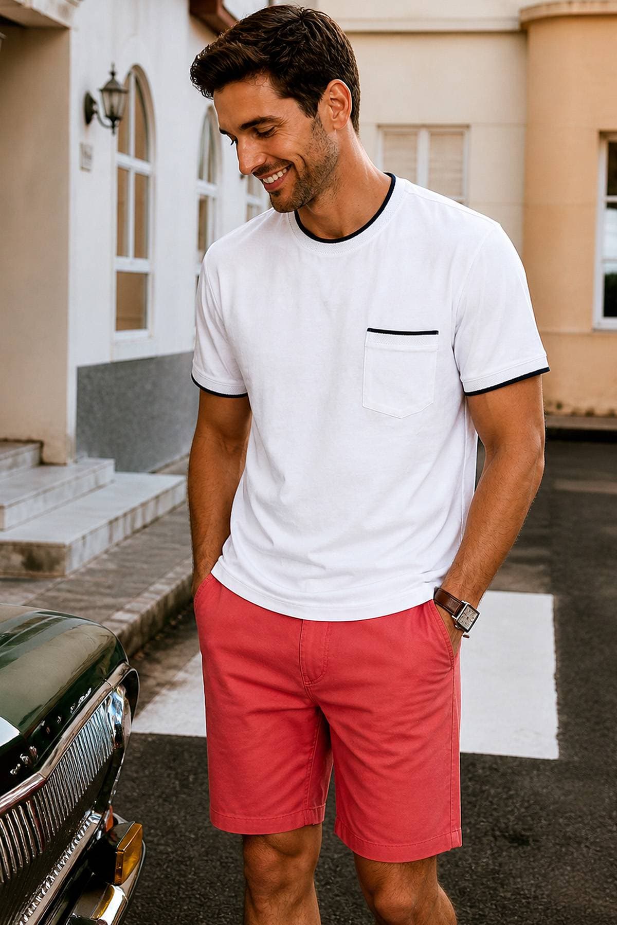 A man is wearing a white T-shirt and pink shorts, smiling next to a vintage car.