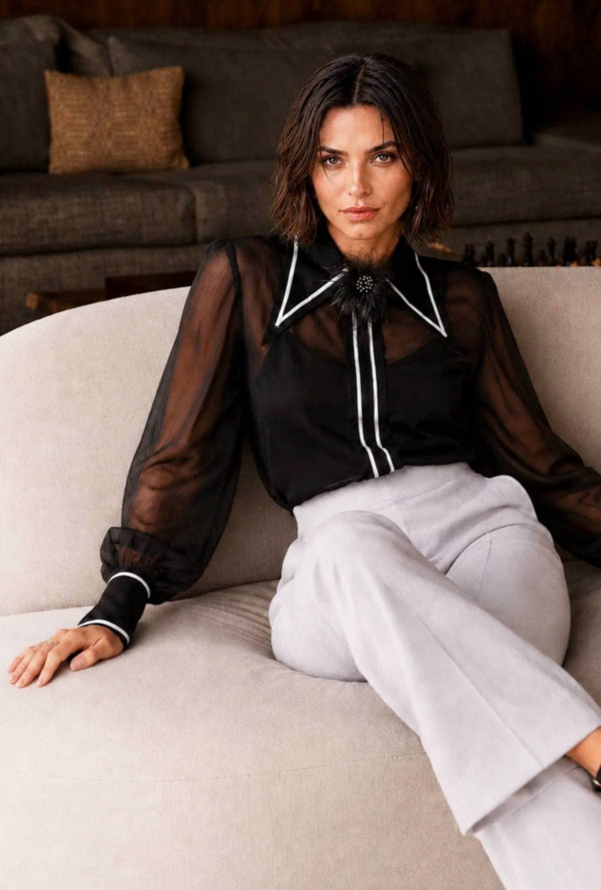 A person wearing a sheer black blouse with white detailing and light-colored trousers sits on a light sofa.