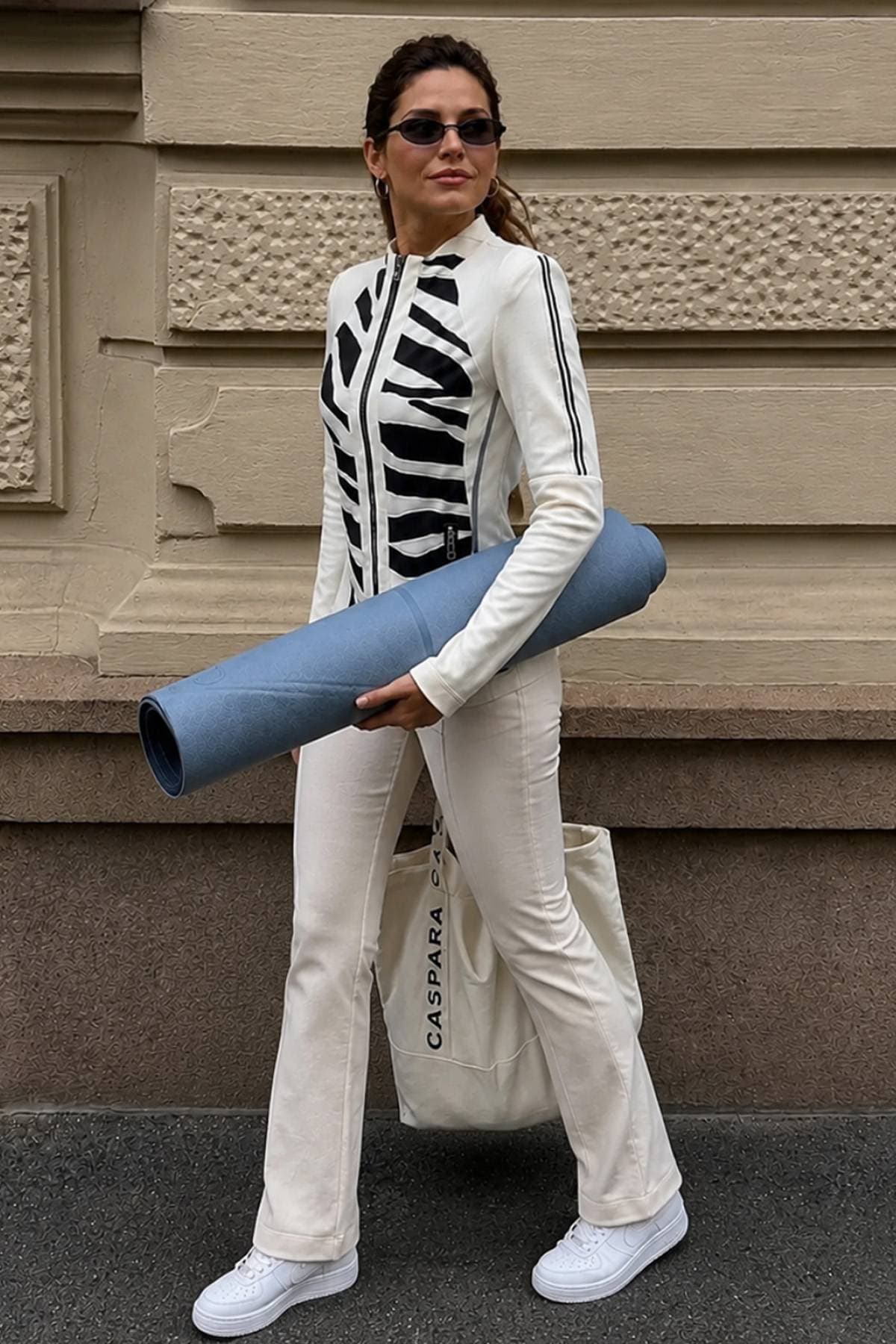 A woman wearing a white and black outfit carries a blue yoga mat and a tote bag.