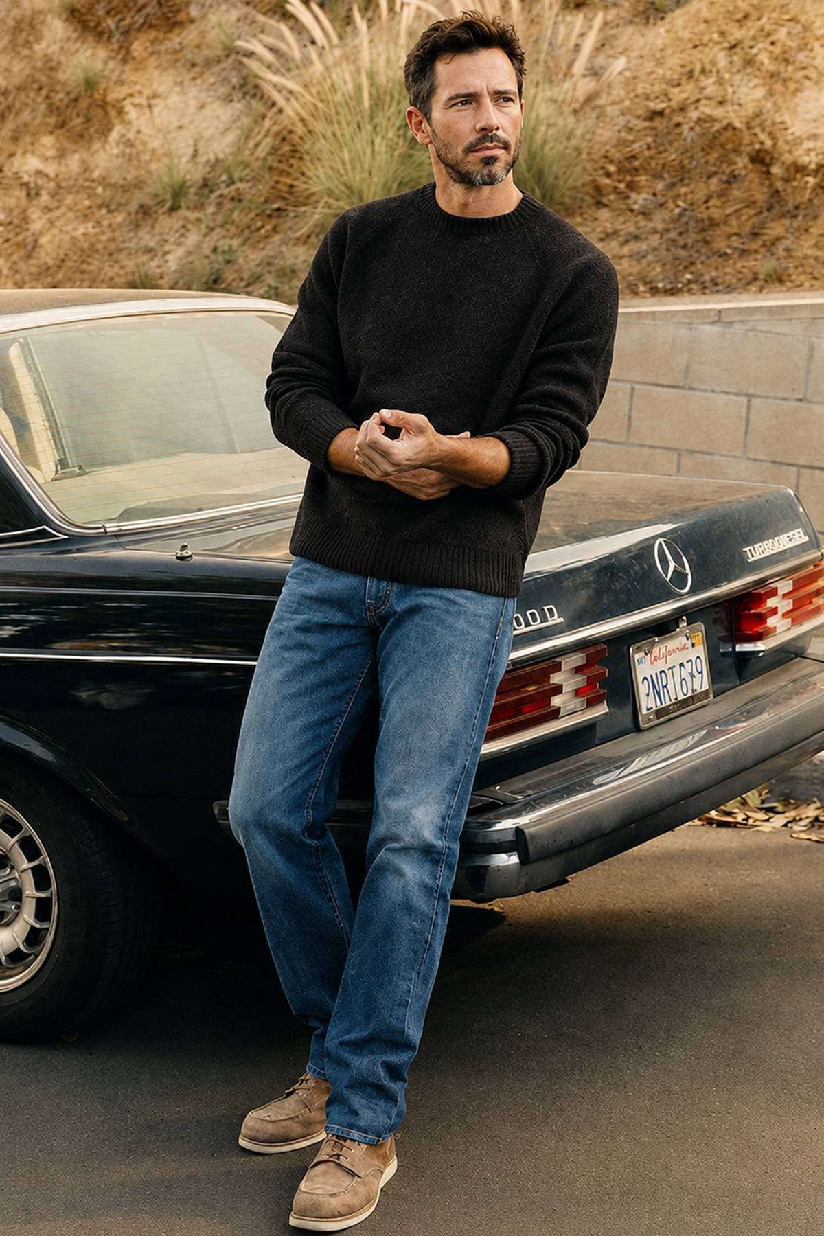 A man in a black sweater and jeans leans against the back of a classic Mercedes car.