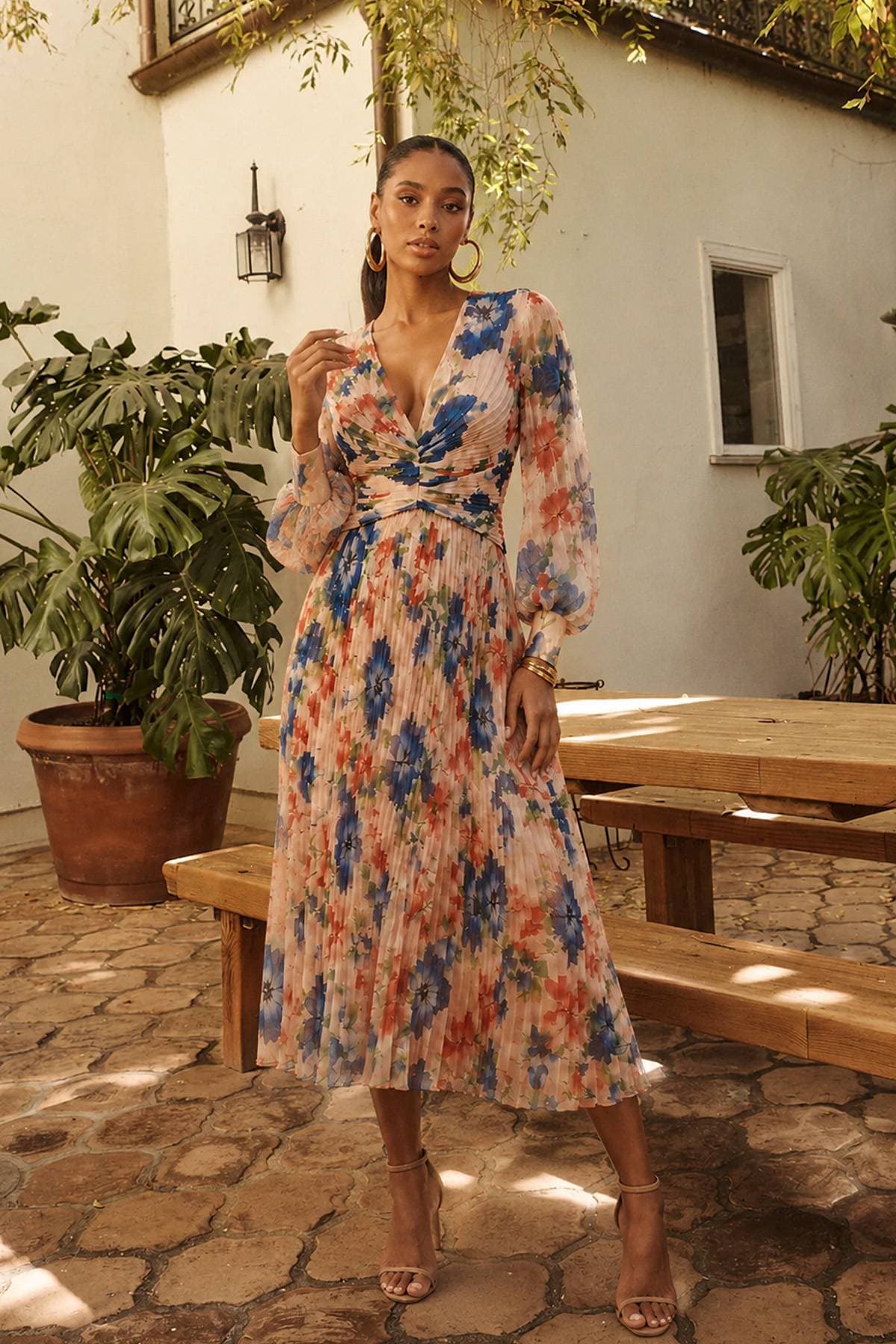 A woman wearing a floral dress poses confidently with her hand raised near her face.
