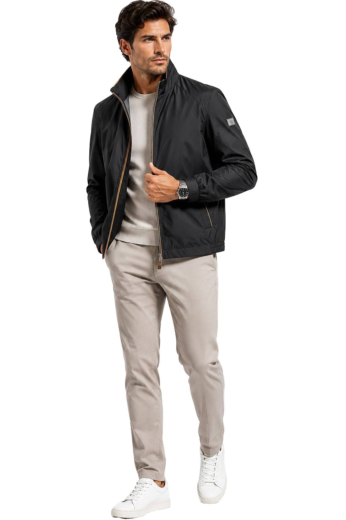 A man is wearing a black jacket over a beige sweater, light khaki pants, and white sneakers.