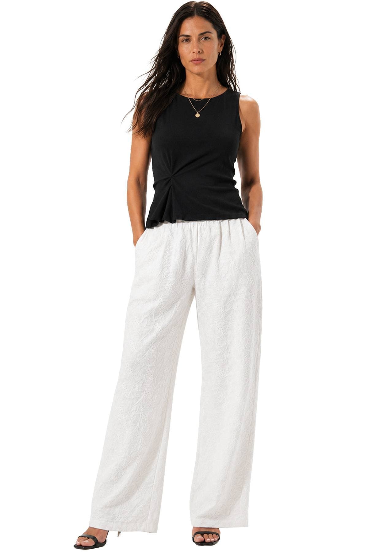 A woman stands wearing a black sleeveless top and white wide-leg pants with her hands in her pockets.