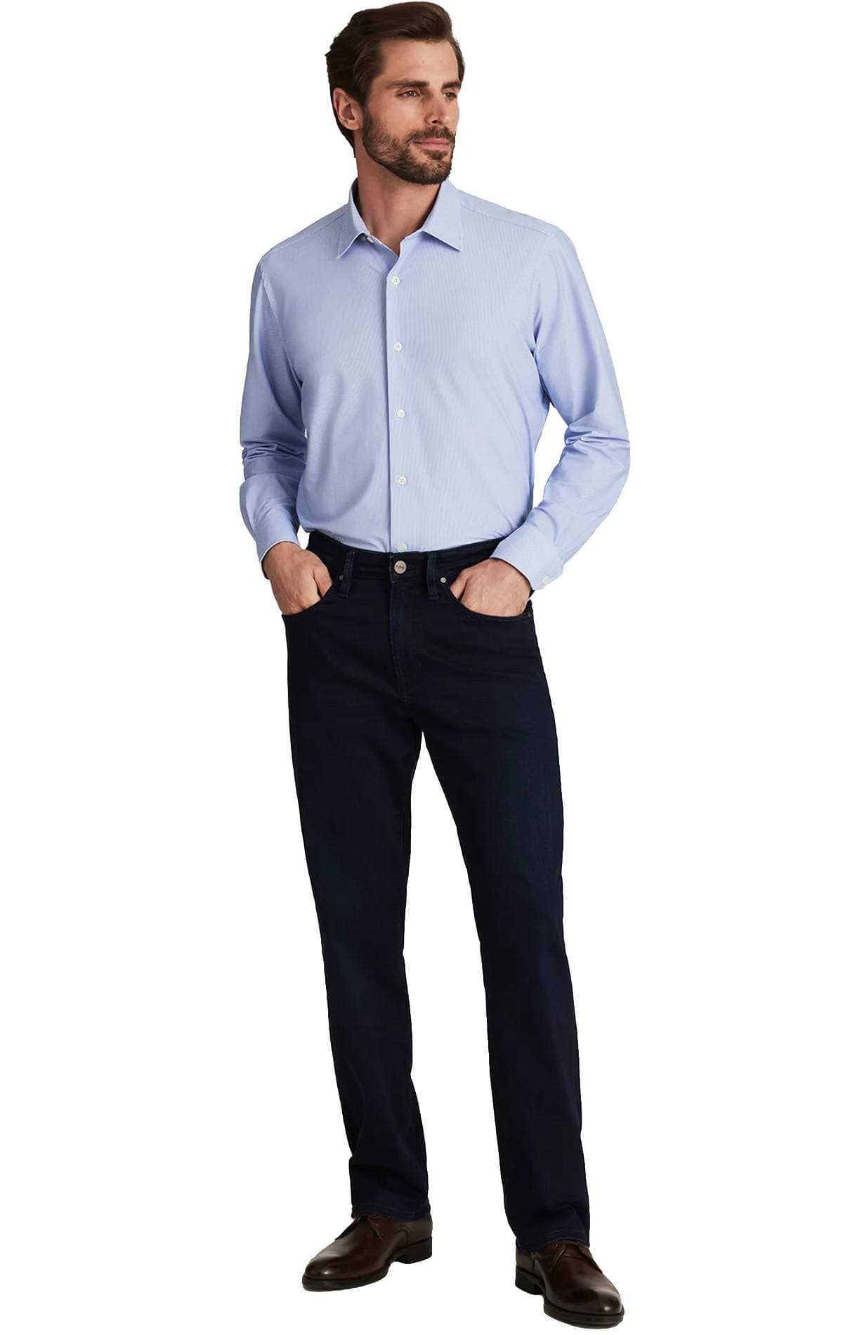 A man in a light blue dress shirt and dark jeans stands with hands in pockets.