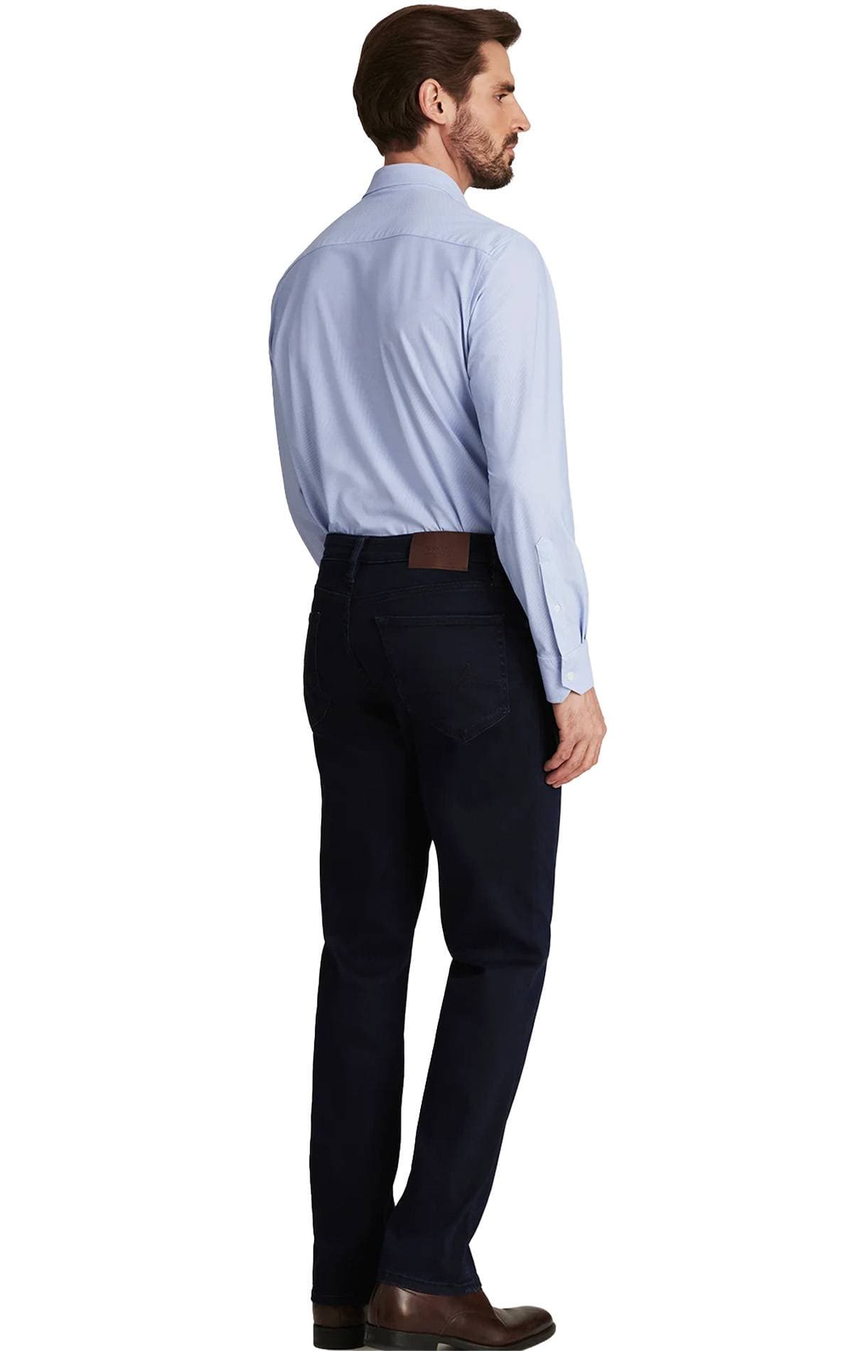 A man wearing a light blue shirt and dark jeans is standing and looking to the side.
