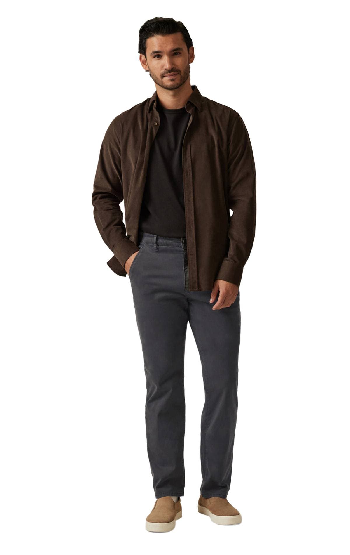 A man stands wearing a brown jacket, black shirt, dark pants, and brown slip-on shoes.