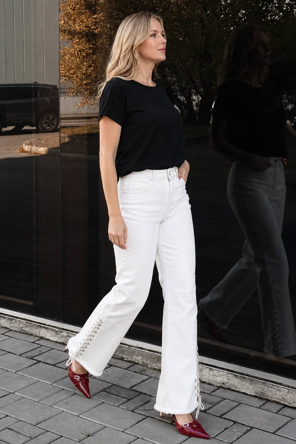 A woman wearing a black shirt and white pants walks confidently while reflecting in a glass wall.