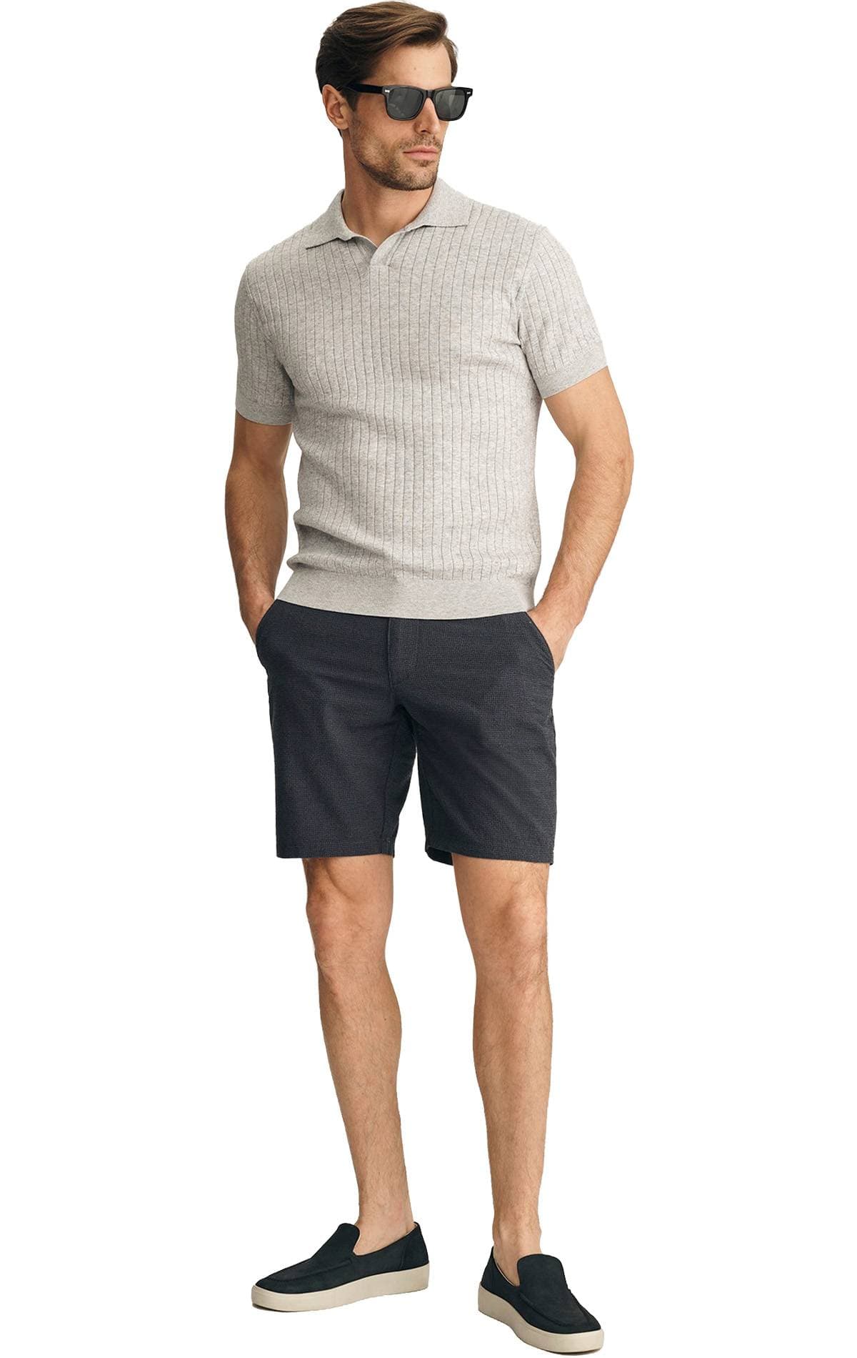 A man wearing a gray ribbed short-sleeve polo shirt, dark shorts, sunglasses, and slip-on shoes stands with hands in pockets.