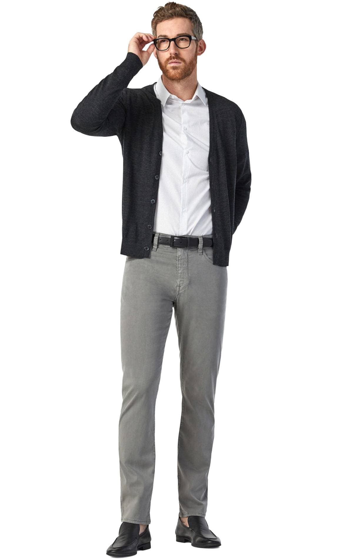 A man in a white shirt, dark cardigan, grey pants, and black shoes adjusts his glasses.